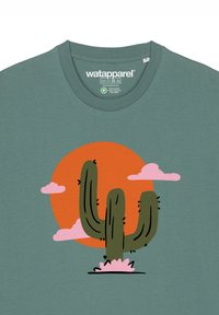 Groene katoenen t-shirt met een grafisch ontwerp van een cactus tegen een oranje cirkel en roze wolken. Het ontwerp is simpel en kleurrijk.