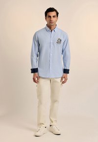 Camisa de manga larga a rayas en azul claro con cuello abotonado, acentos en azul marino en los puños y logo bordado. Pantalones cargo crema y zapatillas blancas.