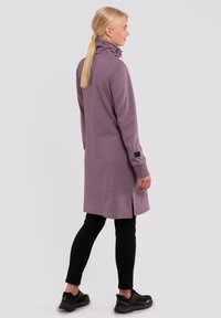 Langärmliges, lavendelfarbenes Pulloverkleid mit hohem Kragen, seitlichen Schlitzen und einem schwarzen Patch am Ärmel, kombiniert mit schwarzen Leggings und Schuhen.