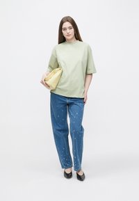 Lichtgroene oversized blouse, blauwe gerafelde spijkerbroek en een gele clutch. Zwarte hakken maken de look compleet. Effen achtergrond.