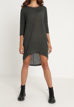 Femme portant une robe ample gris foncé à coupe asymétrique, manches trois-quarts, et des bottines noires à lacets, debout jambes écartées.