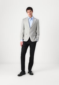 Lichtgrijze blazer met notch-lapels, twee knopen en een borstzak, gecombineerd met een lichtblauw shirt en zwarte broek. Zwarte schoenen maken de look af.