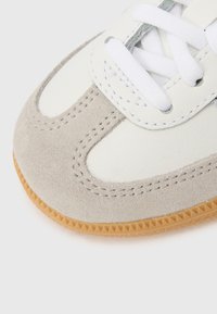 Αθλητικό παπούτσι από λευκό δέρμα με γκρίζες σουέντ λεπτομέρειες, στρογγυλή μύτη και υφασμένο καουτσούκ σόλα σε φυσικό χρώμα. Διαθέτει λευκά κορδόνια και ραφές λεπτομέρειας.