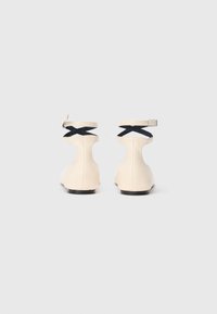 Une paire de chaussures plates beiges avec des brides croisée noires à la cheville et des boucles, présentée de dos sur un fond blanc uni.