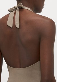 Halter-neck baddräkt i mjuk taupe-tyg, med en knuten rosett på ryggen och en djup skärning i ryggen, slät textur.