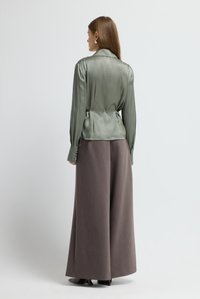 Satin olivgrüne Bluse mit strukturierten Schultern, tailliert und mit geknöpften Manschetten, kombiniert mit lockeren, grau-braunen, weit geschnittenen Hosen.