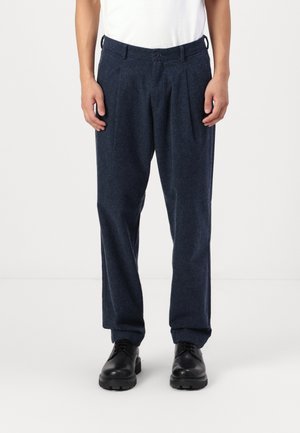 Mężczyzna w ciemnoniebieskich plisowanych spodniach, czarnych skórzanych butach i białej koszuli, stojący na tle jednolitego jasnego tła.