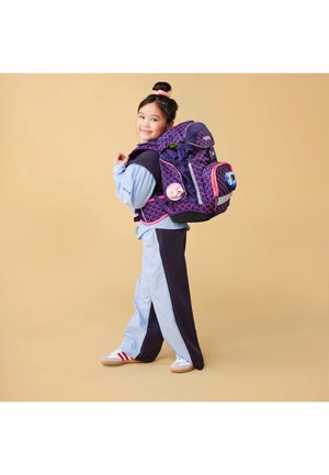 Ragazza sorridente, indossa un outfit azzurro chiaro e blu navy con sneakers bianche, porta uno zaino grande viola e rosa con toppe decorative.