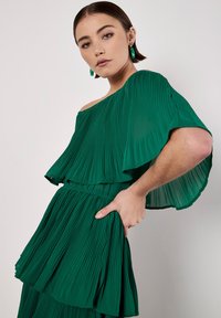 Grünes plissiertes Kleid mit einem-schultrigem Design, geschichteten Rüschengen und gerafftem Taillenbund. Glatte Textur und fließende Silhouette.