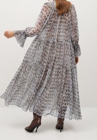 Nainen, joka on pukeutunut pitkään, virtaavaan kuviolliseen mekkoon, jossa on kerroksellisia yksityiskohtia ja levenevät hihansuut, yhdistettynä ruskeisiin korkokenkiin, seisoo sileää taustaa vasten.