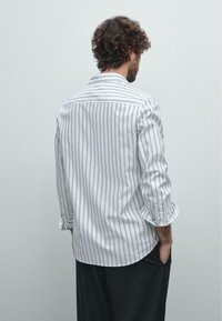 Mann mit lockigen Haaren, der ein weißes Hemd mit dünnen vertikalen grauen Streifen und schwarze Hosen trägt, mit den Händen in den Taschen stehend, von hinten ausgerichtet.