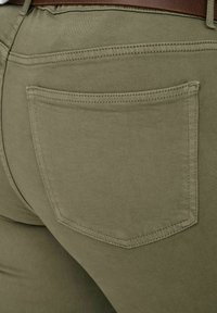 Primer plano del bolsillo de un pantalón verde oliva con costuras visibles y un cinturón marrón en la cintura.