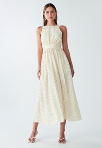 Žena nosi bezriječni kremasti maksi haljinu s crnim točkicama, uparenu s bijelim sandalama na visoku petu i bisernim naušnicama.