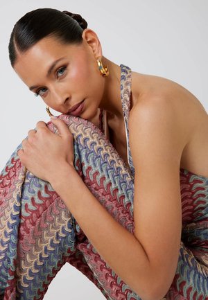 Mehrfarbiger Strickstoff mit einem Wellenmuster in Pink, Blau und Grün, der um einen Arm drapiert ist, dazu glänzende goldene Creolen.