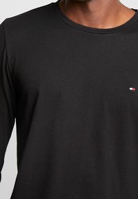 Schwarzes Langarm-T-Shirt aus weichem Stoff. Verfügt über einen runden Ausschnitt und ein kleines Logo mit roten, weißen und blauen Akzenten auf der Brust.