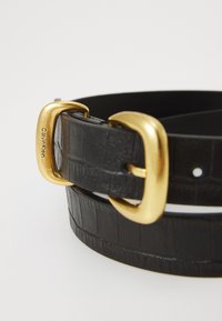 Cintura in pelle nera con texture coccodrillo, dotata di una fibbia in metallo dorato con marchio. Accentuata da una striscia bianca lungo il bordo.