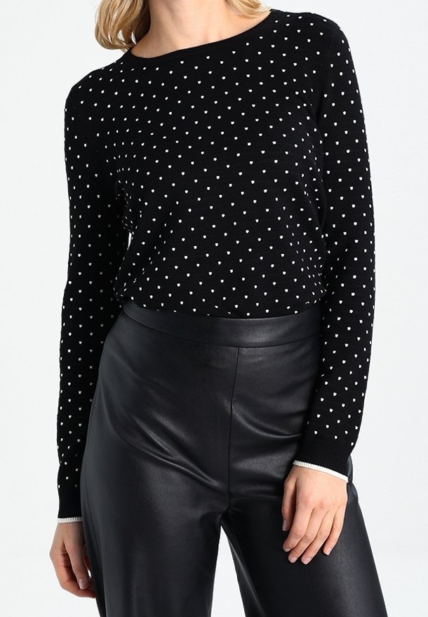 Femme portant un pull noir à manches longues avec de petits cœurs blancs, associé à un pantalon en cuir noir taille haute.
