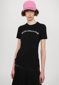 Melna īsām piedurknēm T-kreklu ar uzrakstu "Versace Jeans Couture" izšūtu baltā krāsā. Kombinēts ar rozā cepuri un melnu svārku ar krokām.