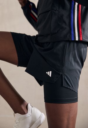 Osoba ubrana w czarne sportowe szorty z warstwowym wzorem, czarną kurtkę z czerwonymi, białymi i niebieskimi paskami oraz białe trampki, w trakcie kroku.