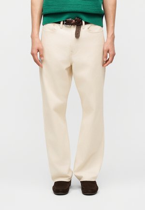 Homme portant un pantalon droit ivoire, une ceinture tressée marron, des chaussures marron foncé et un haut en maille vert à manches courtes, debout les mains détendues.