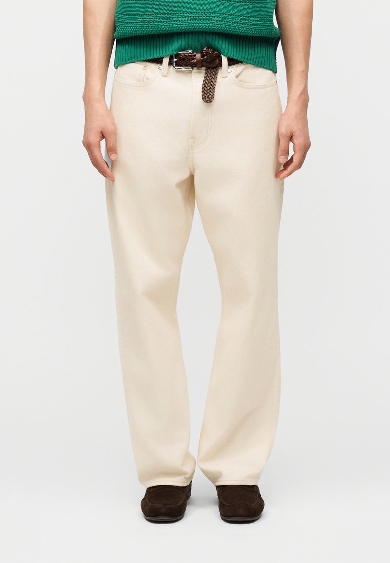 Άνδρας που φοράει πανομοιότυπα παντελόνια εκρού με ίσια γραμμή, καφέ πλεκτή ζώνη, σκούρα καφέ παπούτσια και πράσινο πλεκτό τοπ με κοντά μανίκια, στέκεται με χαλαρά τα χέρια.