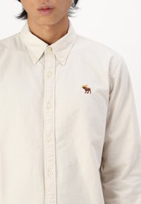 Lys beige knap-op skjorte i bomuld, med en lille brun elg broderi på venstre bryst og klassisk knap-ned krave.