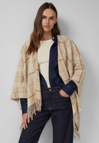 Beżowy poncho w kratę z frędzlami, wykonane z miękkiego materiału, nałożone na granatową koszulę dżinsową i jasną bluzkę.