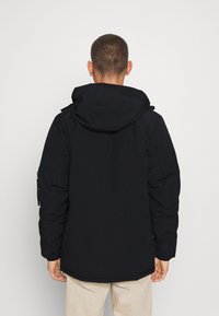 Svart isolerad jacka med huva, med en slät textur, elastiska muddar och en något längre bakhel. Inga synliga logotyper eller utsmyckningar.