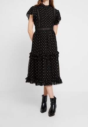 Frau trägt ein schwarzes Midikleid mit kleinen kreisförmigen Mustern, Rüschen an den Ärmeln und am Saum, kombiniert mit schwarzen Stiefeletten, steht mit den Händen hinter dem Rücken.