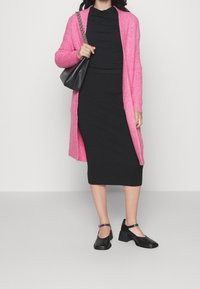 Kvinna i en lång rosa kcardigan, svart topp och kjol, svarta blockklackade Mary Jane-skor, som håller i en svart handväska med chain strap.