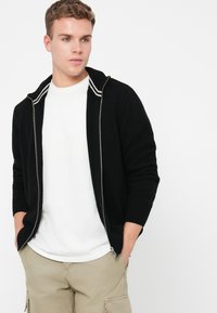 Nuori mies, jolla on lyhyet hiukset, pukeutuneena mustaan vetoketjulliseen villapaitaan, jossa on valkoisia raitoja, ja valkoiseen t-paitaan, vaaleisiin cargo-housuihin, kädet taskuissa.