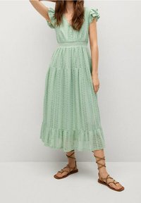 Robe maxi vert menthe avec un motif texturé, des manches volantées, un détail de boutons au niveau du décolleté et un ourlet évasé. Portée avec des sandales à lanières marron.