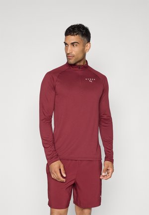 Top sportivo a maniche lunghe color bordeaux con colletto alto e mezza zip, dotato di un logo sul petto, abbinato a pantaloni corti coordinati. Tessuto con texture liscia.