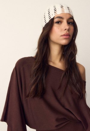 Mladá žena s dlhými tmavými vlasmi má na hlave bielu pletenú čiapku a hnedý sveter s odhalenými ramenami, hľadí mierne do strany na jednoduchom pozadí.