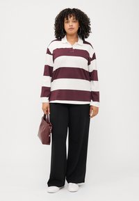 Une personne aux cheveux bouclés se tient avec confiance, portant une chemise à rayures à manches longues, un pantalon noir et tenant un sac à main bordeaux.
