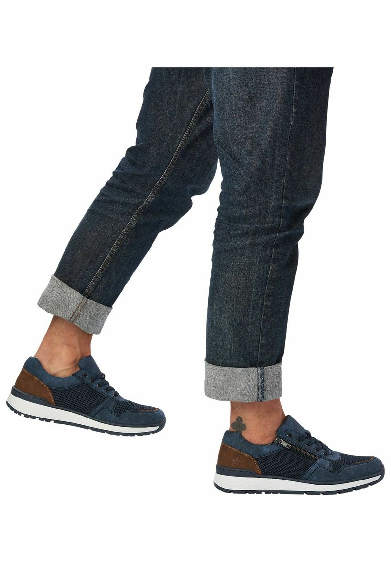 Baskets de sport bleu marine avec des accents en cuir marron, des panneaux en mesh, des semelles blanches et des bandes en caoutchouc noir, associées à un pantalon en denim foncé retroussé.