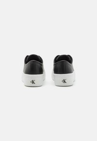 Sorte skinnesko med hvit såle, bakperspektiv som viser avrundet design og logo detaljer på hælen. Glatt tekstur, minimalistisch stil.