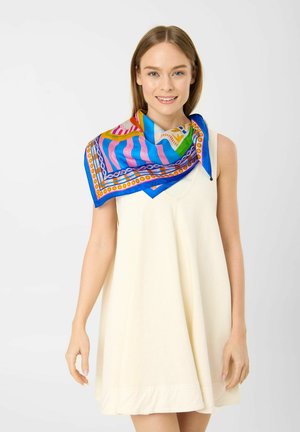 Junge Frau lächelt, trägt ein ärmelloses cremefarbenes Kleid und einen bunten, gemusterten Schal, der über eine Schulter und um den Hals drapiert ist.