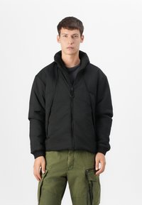 Veste noire zippée en tissu texturé, avec col montant et poignets élastiques, associée à un pantalon cargo vert avec poches latérales et zips.