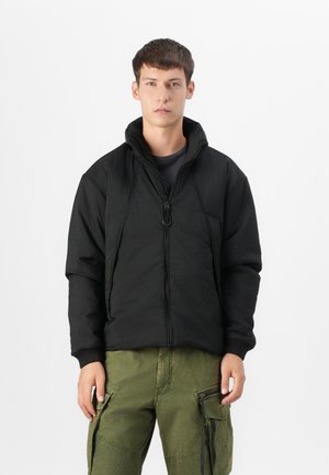 Veste noire zippée en tissu texturé, avec col montant et poignets élastiques, associée à un pantalon cargo vert avec poches latérales et zips.
