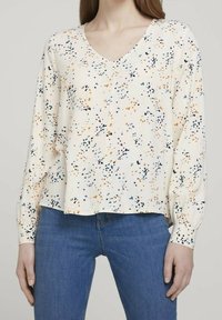 Cremfarbene Bluse mit V-Ausschnitt, langen Ärmeln und lässiger Passform, die ein farbenfrohes Spritzmuster in Orange, Blau und Schwarz aufweist.
