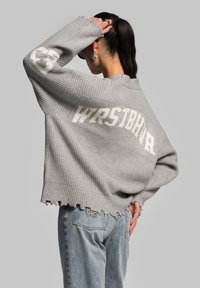 Gray knitted sweater with oversized fit, featuring the text "WRSTBHVR" in white on the back. Frayed edges at the sleeves and bottom.