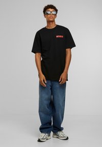 Sort oversized t-shirt med rød "DAYS BEFORE" tekst, parret med løse blå jeans og grå sneakers med sølv accenter.