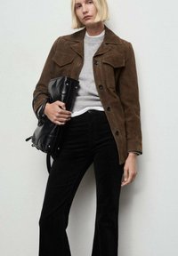 Mujer rubia vestida con una chaqueta de ante marrón, suéter gris, pantalones negros, sosteniendo un gran bolso de cuero negro contra una pared clara y simple.
