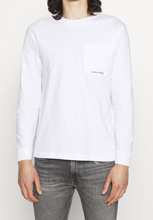 Personne portant une chemise blanche à manches longues avec une poche poitrine portant l'inscription "Calvin Klein Jeans" et un jean gris délavé.
