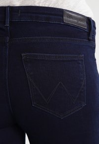 Mörkblå denimjeans med en bakficka som har en distinkt zigzag-stygn. Midjebandet har en svart läderlogopatch.