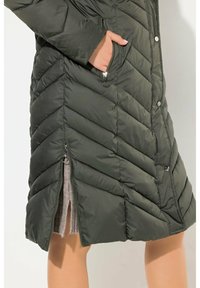 Olivgrön puffjacka med chevron-quilting, tryckknappar, sidofickor och dragkedjeöppning i botten, som har ett mjukt grått innerfoder.
