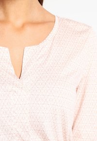 Lichtroze blouse met een V-hals, gestructureerde stof met een subtiel bloemenpatroon en een losse pasvorm die de schouders accentueert.