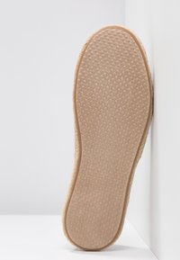 Semelle d'une chaussure à enfiler marron clair avec une semelle en caoutchouc texturée, posée debout contre un mur d'angle blanc.