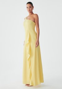 Yellow, one-shoulder maxi dress with a textured fabric, featuring a loose, ruffled panel down the front and a flowing silhouette.